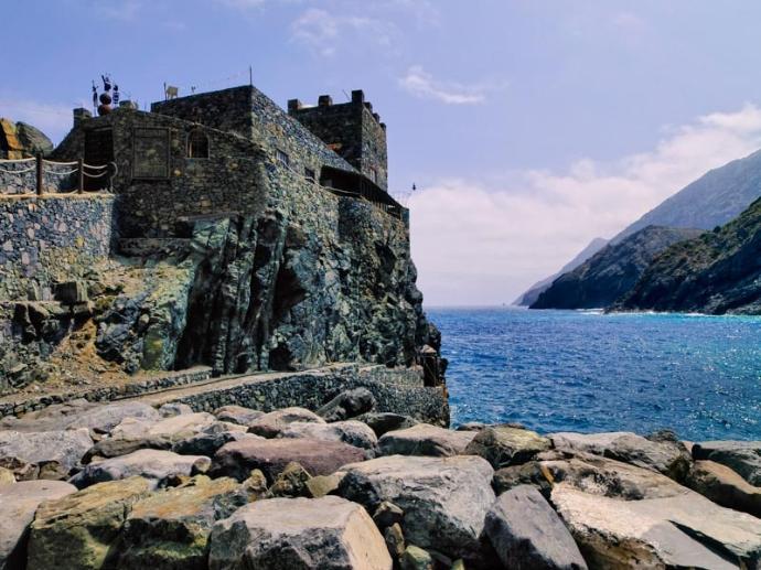 Se ve al Castillo del mar en el borde dando al mar, y un camino de piedras y sendero para llegar a él, con montañas de fondo por el otro lado